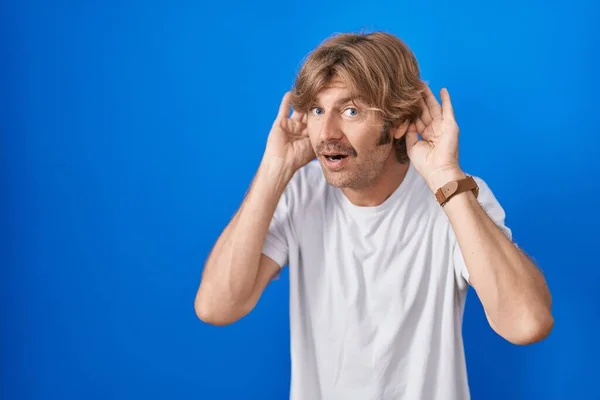 中年の男性の青い背景に耳のジェスチャーで両方の手を聞いて ゴシップを好奇心をしようと立っている 聴覚障害や聴覚障害 — ストック写真
