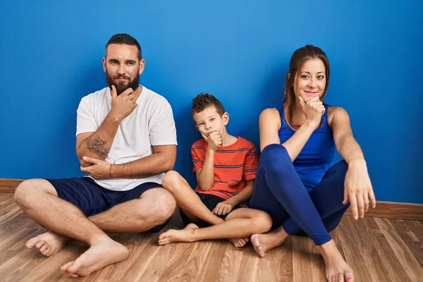 Family of three sitting on the floor at home looking confident at the camera with smile with crossed arms and hand raised on chin. thinking positive.