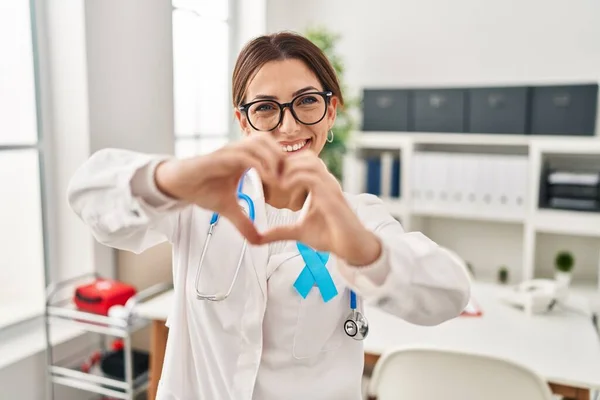 Young brunette doctor woman wearing stethoscope at the clinic smiling in love doing heart symbol shape with hands. romantic concept.