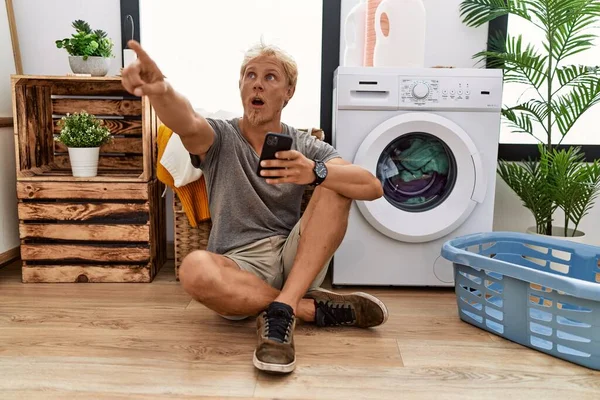Young Blond Man Doing Laundry Using Smartphone Pointing Finger Surprised — Stok fotoğraf