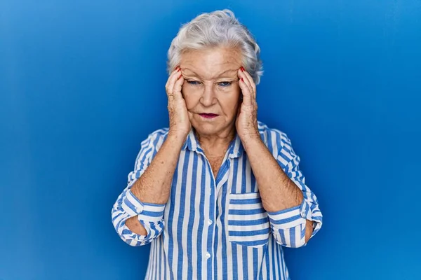 ストレスが原因で頭の痛みのために頭の上に手で青の背景に立って灰色の髪を持つシニア女性 片頭痛 — ストック写真