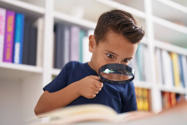Adorable hispanic toddler student reading book using magnifying glass at library school