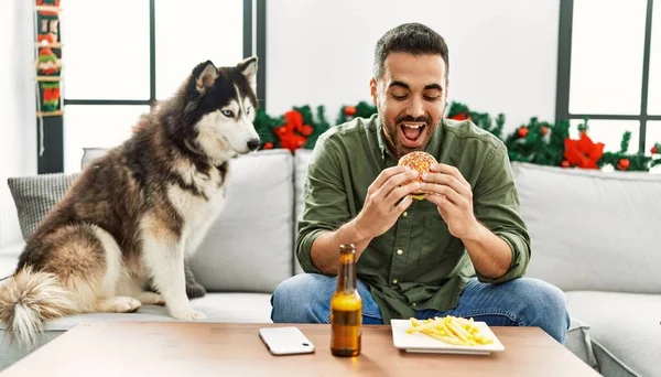 Young hispanic man eating hamburger sitting on sofa with dog by christmas decor at home