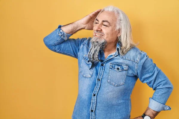 Middle age man with grey hair standing over yellow background smiling confident touching hair with hand up gesture, posing attractive and fashionable