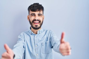 Young hispanic man with beard standing over blue background approving doing positive gesture with hand, thumbs up smiling and happy for success. winner gesture. 