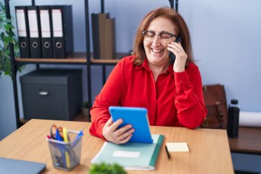 Senior woman business worker using touchpad talking on smartphone at office