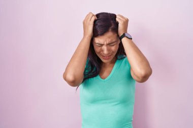 Pembe arka planda duran genç İspanyol kadın acı ve migren yüzünden çaresiz ve stresli baş ağrısı çekiyor. Eller başının üstünde.. 