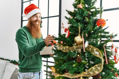 Genç kızıl saçlı adam gülümsüyor. Evde kendine güvenen bir Noel ağacı süslüyordu.