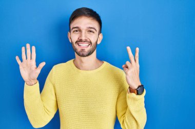 Hispanic man standing over blue background showing and pointing up with fingers number seven while smiling confident and happy. 