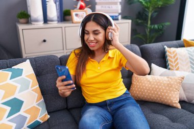 Young beautiful arab woman listening to music sitting on sofa at home