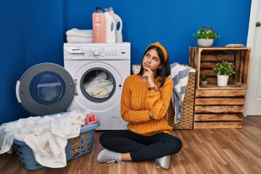 Young hispanic woman doing laundry with hand on chin thinking about question, pensive expression. smiling with thoughtful face. doubt concept. 