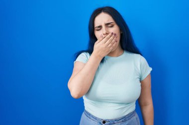 Young modern girl with blue hair standing over blue background bored yawning tired covering mouth with hand. restless and sleepiness. 