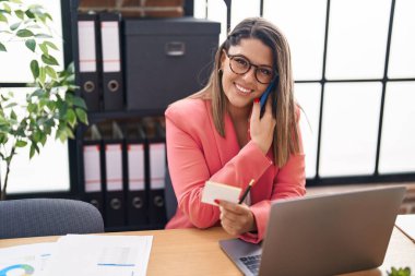 Young hispanic woman business worker talking on smartphone reading reminder paper at office