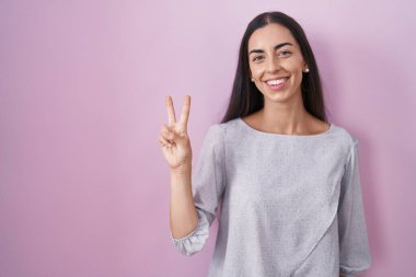 Genç esmer kadın pembe arka planda duruyor. İki numarayı gösterip işaret ediyor. Kendinden emin ve mutlu bir şekilde gülümsüyor.. 