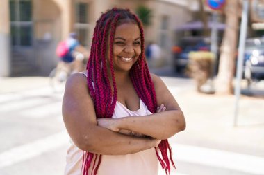 Afro-Amerikalı kadın gülümsüyor. Kollarını kavuşturup caddede duruyor.