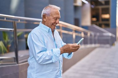 Yaşlı, gri saçlı, kendine güvenen, caddede akıllı telefon kullanan bir adam.