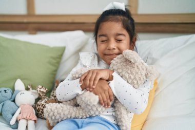 Adorable hispanic girl hugging teddy bear lying on bed at bedroom