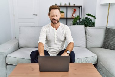 Middle age man using laptop at home winking looking at the camera with sexy expression, cheerful and happy face. 