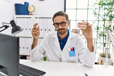 Young hispanic dentist man holding denture and toothbrush relaxed with serious expression on face. simple and natural looking at the camera. 