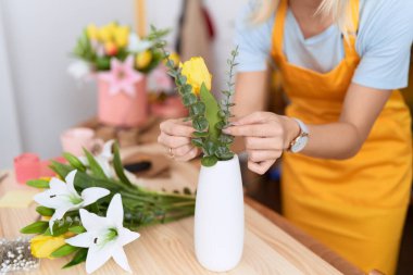 Young blonde woman florist putting flowers on jar at flower shop