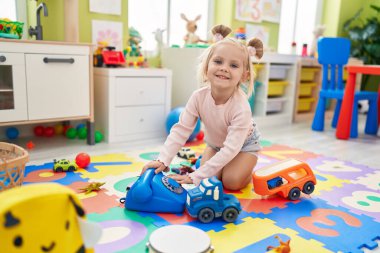 Anaokulunda yerde oturan beyaz bir kız telefon oyuncağıyla oynuyor.