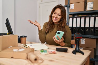 Hispanic woman working at small business using smartphone celebrating achievement with happy smile and winner expression with raised hand 
