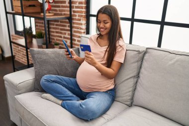 Genç Latin bir kadın hamile. Evde akıllı telefon ve kredi kartı kullanıyor.