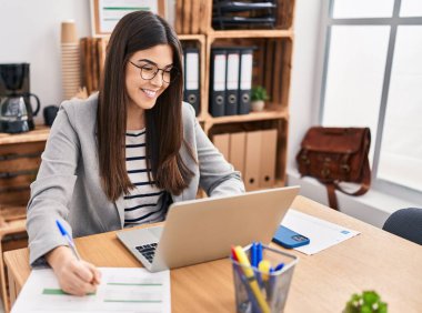 Genç, güzel İspanyol iş kadını. Ofisteki evraklarda dizüstü bilgisayar kullanıyor.