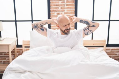 Genç kel adam yatak odasında kollarını esneterek uyanıyor.
