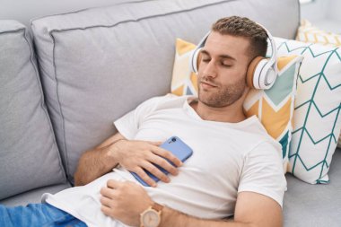 Young caucasian man listening to music lying on sofa at home