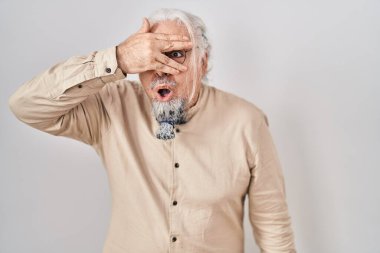 Middle age man with grey hair standing over isolated background peeking in shock covering face and eyes with hand, looking through fingers with embarrassed expression. 