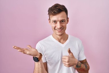 Pembe arka planda duran beyaz bir adam avuç içini gösteriyor ve baş parmaklarıyla iyi bir hareket yapıyor, mutlu ve neşeli gülümsüyor. 