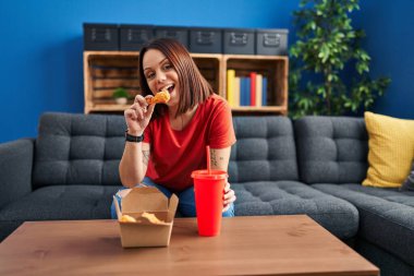 Young beautiful hispanic woman eating fried chicken sitting on sofa at home
