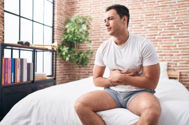 Young hispanic man suffering for stomach ache sitting on bed at bedroom