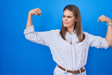 Hispanic young woman standing over blue background showing arms muscles smiling proud. fitness concept. 