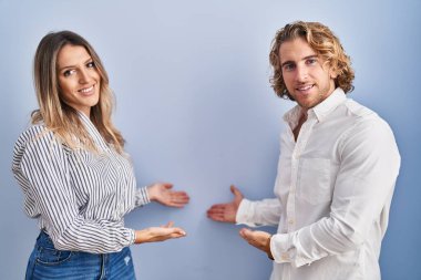 Young couple standing over blue background inviting to enter smiling natural with open hand 