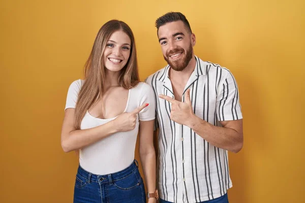Young couple standing over yellow background cheerful with a smile on face pointing with hand and finger up to the side with happy and natural expression