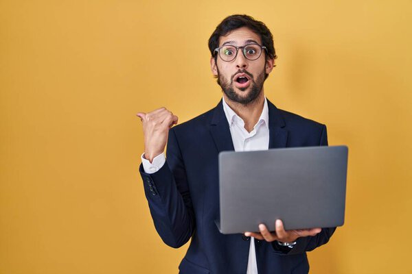 Handsome latin man working using computer laptop surprised pointing with hand finger to the side, open mouth amazed expression. 