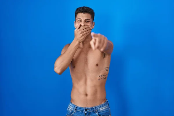 Young hispanic man standing shirtless over blue background laughing at you, pointing finger to the camera with hand over mouth, shame expression