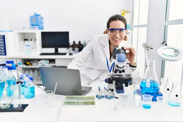 研究室でノートパソコンと顕微鏡を使って科学者の制服を着た若いヒスパニック系の女性 — ストック写真