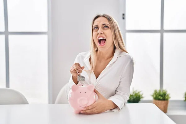 Young Blonde Woman Holding Piggy Bank House Keys Angry Mad — 스톡 사진