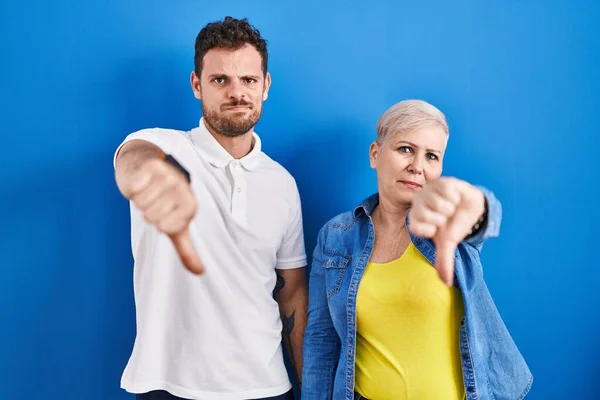 Young brazilian mother and son standing over blue background looking unhappy and angry showing rejection and negative with thumbs down gesture. bad expression.