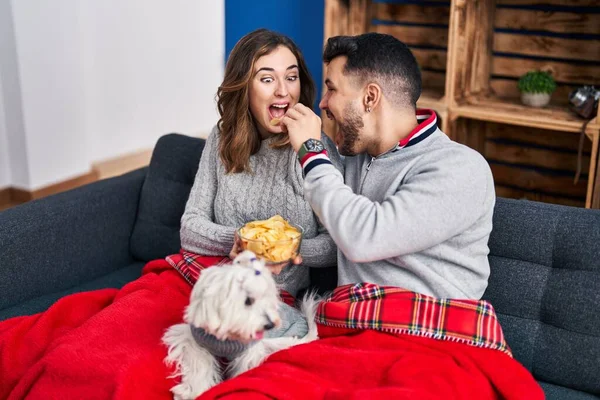 Man and woman eating chips potatoes sitting on sofa with dog at home