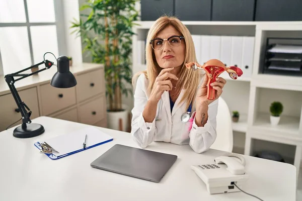 Middle age blonde gynecologist woman holding anatomical model of female genital organ serious face thinking about question with hand on chin, thoughtful about confusing idea