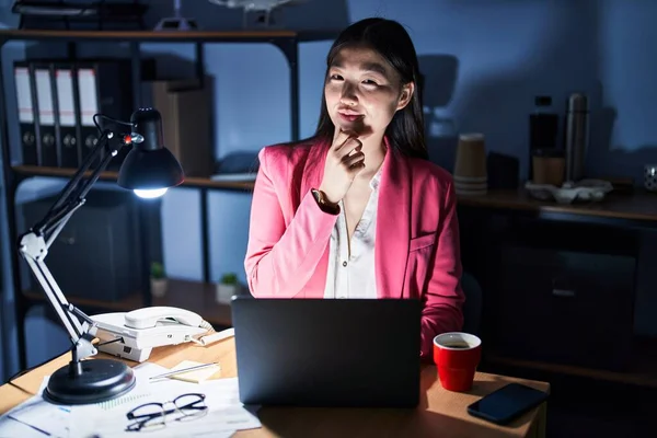 Chinese young woman working at the office at night looking confident at the camera smiling with crossed arms and hand raised on chin. thinking positive.