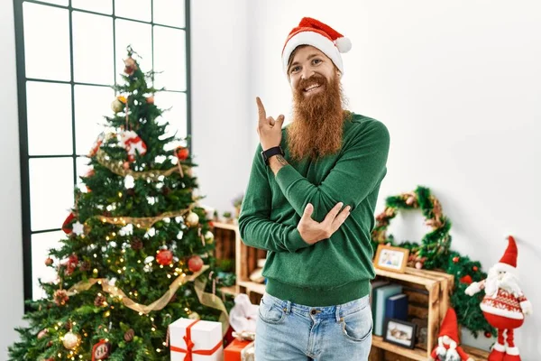 红头发男人留着长胡子 头戴圣诞帽 头戴圣诞礼帽 脸上挂着大大的笑容 手指头指向旁边看相机 — 图库照片