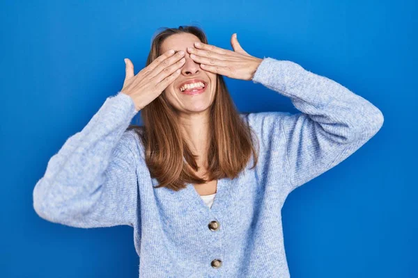 笑顔で陽気で面白い手で目を覆う青い背景の上に立つ若い女性 視覚障害の概念 — ストック写真