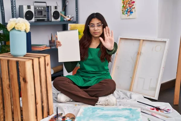 Hispanic young woman holding notebook at art studio with open hand doing stop sign with serious and confident expression, defense gesture