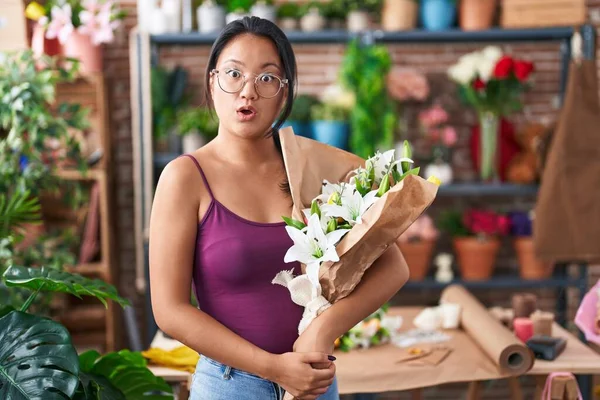 Asian young woman at florist shop holding bouquet of flowers scared and amazed with open mouth for surprise, disbelief face