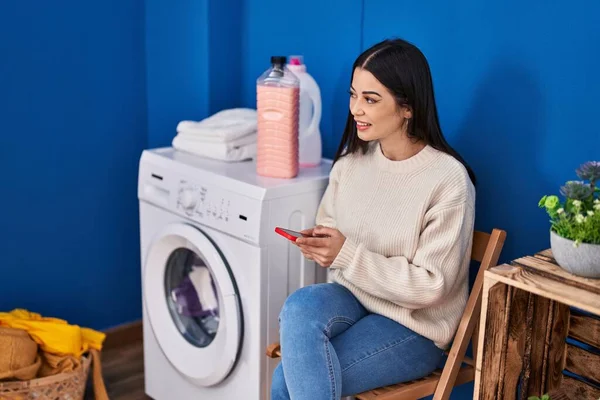 年轻美丽的惊慌失措的女人用智能手机在洗衣房等洗衣机 — 图库照片